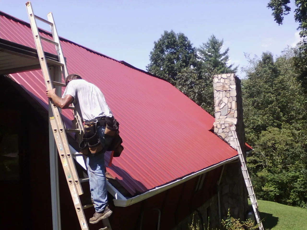 Roofer on a ladder beside a red metal roof and stone chimney — Soffit & Fascia Repair by Roofing Brothers in Topeka
